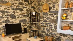 A living room with a stone wall, a wooden TV stand, a black metal table, a grey sofa, and a woven rug. - Jade's House BBQ & Sea View Villa (Calheta)