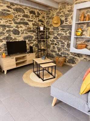 A living room with a stone wall, a wooden TV stand, a black metal table, a grey sofa, and a woven rug.