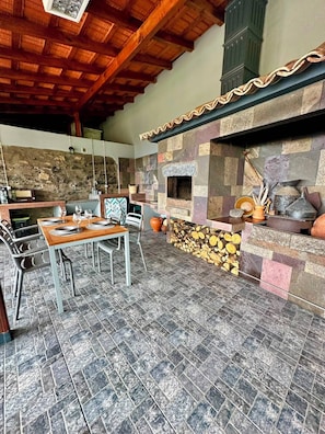 Un espace extérieur aménagé pour les repas, doté d’une cheminée en pierre, d’un plafond en bois et d’un sol carrelé.