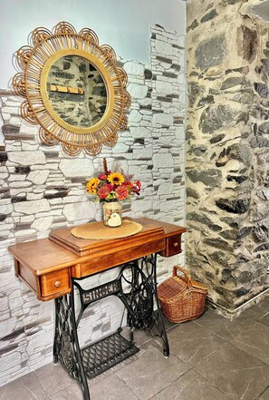 A vintage Singer sewing machine with a wooden table, a decorative mirror, and a vase with flowers against a stone wall backdrop.
