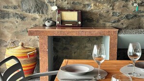 A dining table set with plates, glasses, and cutlery, a chair, a radio, and a stone wall in the background. - Jade's House BBQ & Sea View Villa (Calheta)