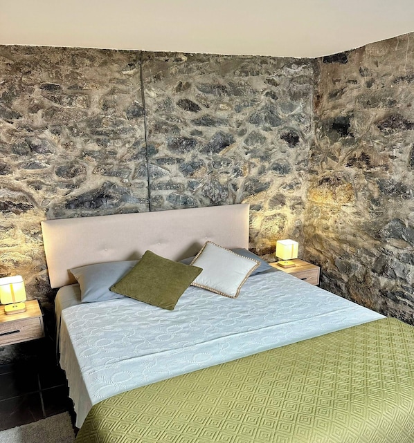 Une chambre avec un mur en pierre, un lit avec une literie à motifs, deux lampes de chevet et un plaid vert.