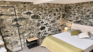 A bedroom with a stone wall, a bed with a white headboard, a bedside table, and a nightstand with a lamp. - Jade's House BBQ & Sea View Villa (Calheta)