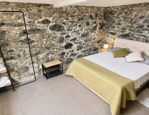 A bedroom with a stone wall, a bed with a white headboard, a bedside table, and a nightstand with a lamp.