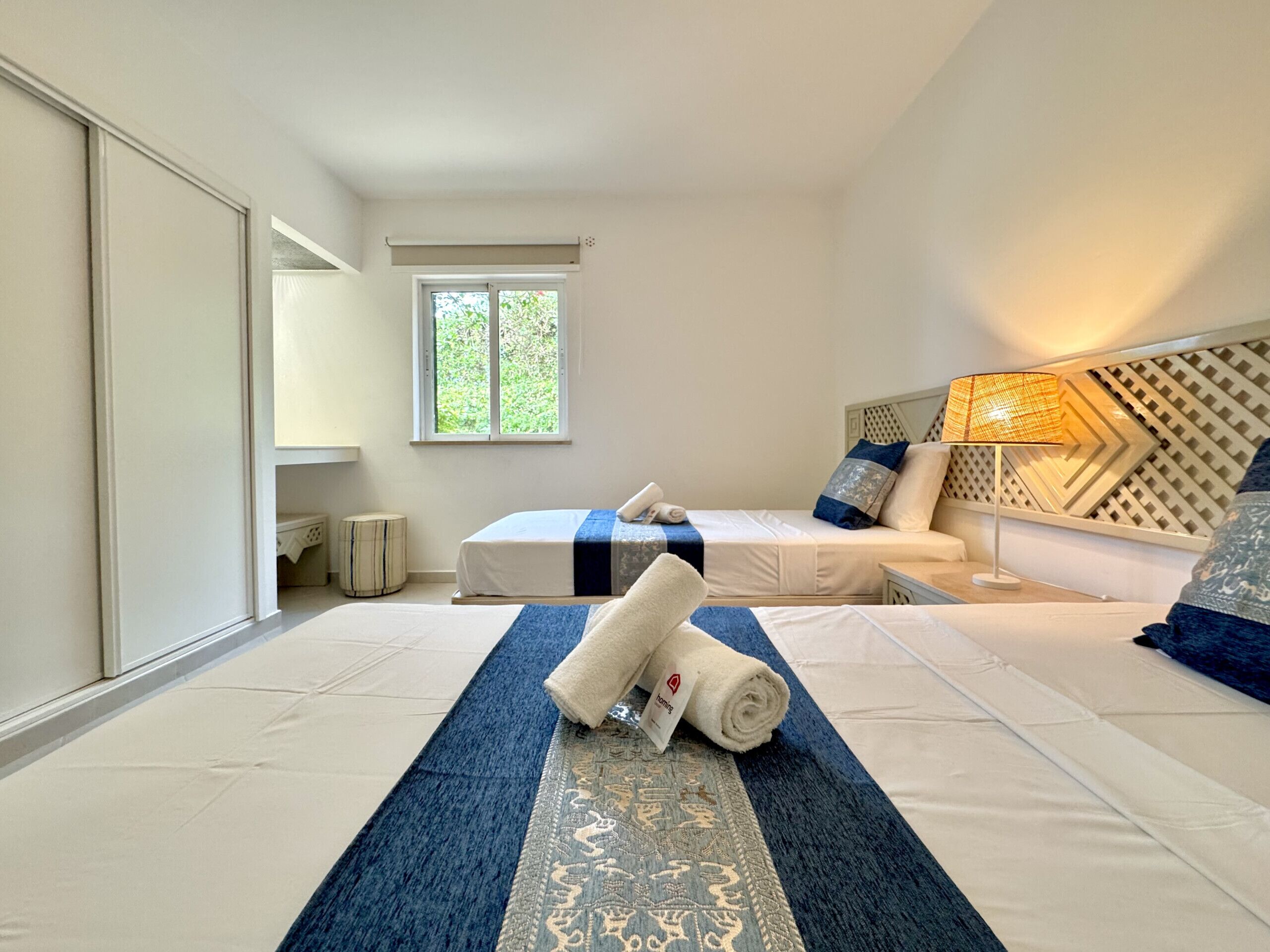 A hotel room with two beds, a window, a lamp, and a patterned wall.