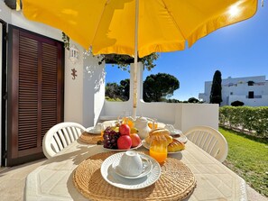 Une table dressée pour le petit-déjeuner, avec un parasol jaune, des chaises blanches et une vue sur un bâtiment blanc et de la verdure.