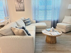 Ein Wohnzimmer mit einem beigefarbenen Ecksofa, einem runden Couchtisch mit Zeitschriften und einem Palmen-Gemälde an der Wand.