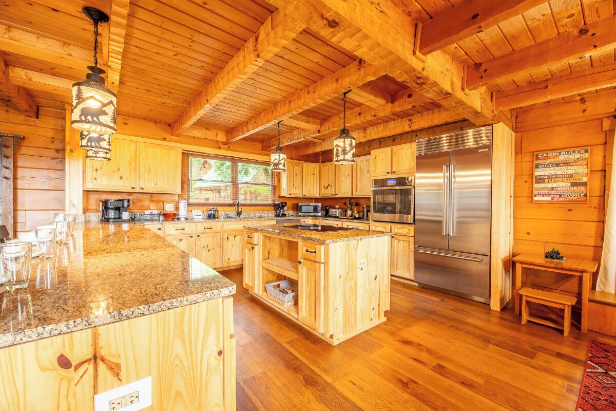 A wooden cabin interior with a kitchen featuring granite countertops, wooden cabinets, stainless steel appliances, and pendant lighting. - Big Bear Lodge - 7BR Mtn Retreat, Theater & Hot Tub (Sevierville)