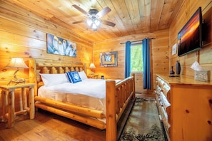 Ein Zimmer in einer Holzhütte mit Bett, Deckenventilator, Fernseher und einem Fenster mit Vorhängen.