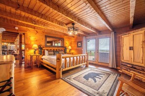 Interieur van een blokhut met een houten bed, een open haard, een eethoek en een groot raam.
