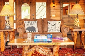 A cozy cabin interior with a wooden couch, two lamps, a table with books and a board game, and a window with a view. - Big Bear Lodge - 7BR Mtn Retreat, Theater & Hot Tub (Sevierville)