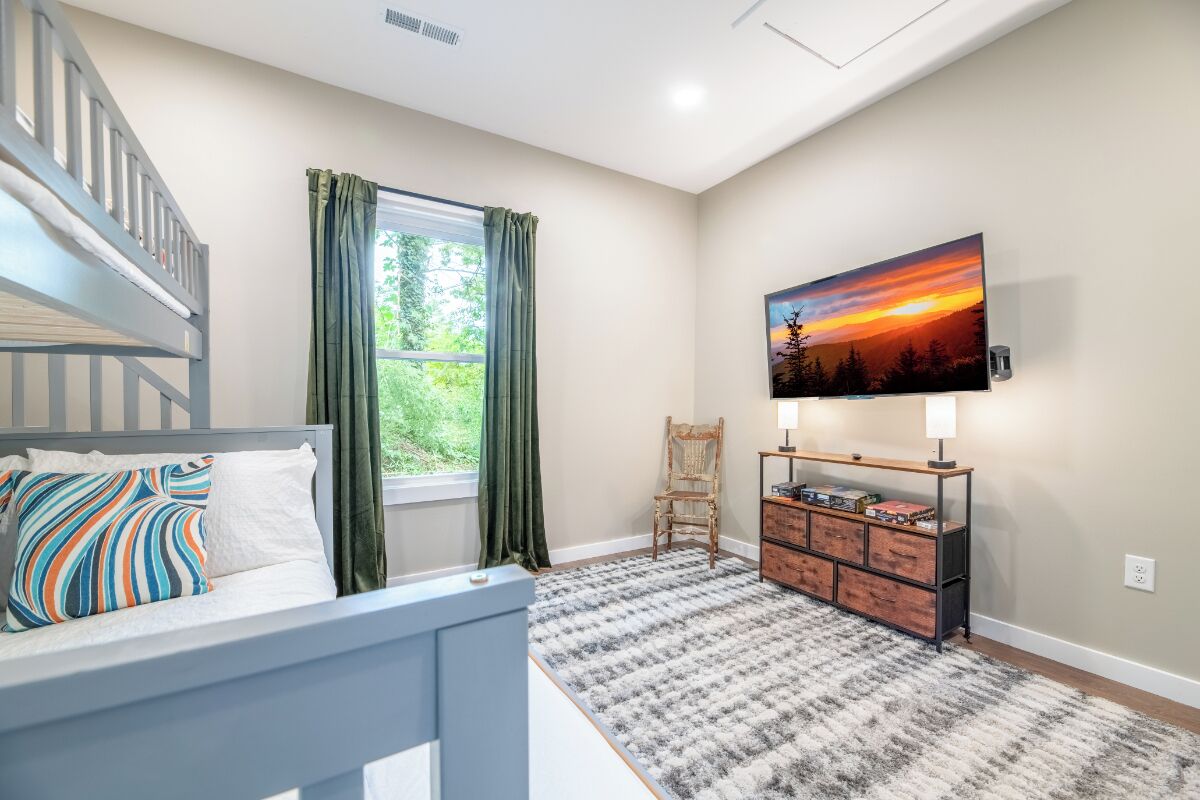 A bedroom with a bunk bed, a window with curtains, a desk with a lamp, a chair, and a TV mounted on the wall.