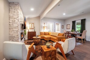 A modern living room with a stone fireplace, leather sofa, wooden coffee table, and a dining area with a table and chairs. - Little River | Hot Tub + Games & Great Location (Walland)