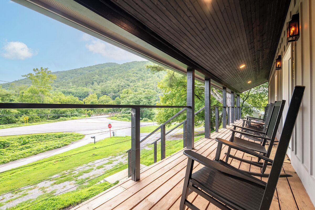 A covered porch with wooden flooring and metal railings, overlooking a green landscape and a road.