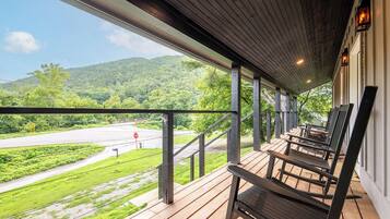 A covered porch with wooden flooring and metal railings, overlooking a green landscape and a road.