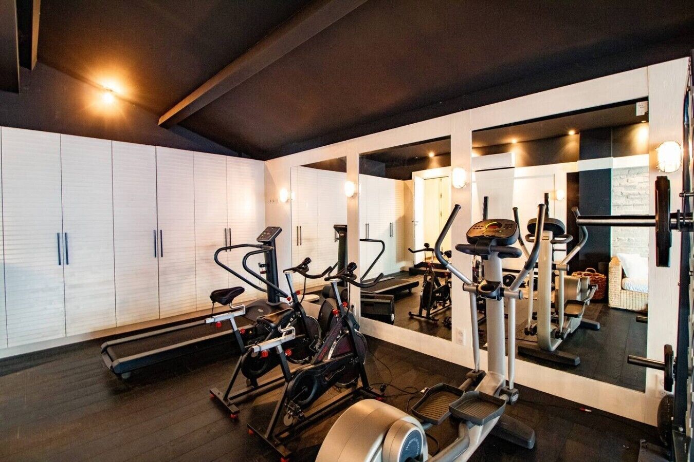 A gym with various exercise equipment, including treadmills and stationary bikes, and a large mirror reflecting the space.