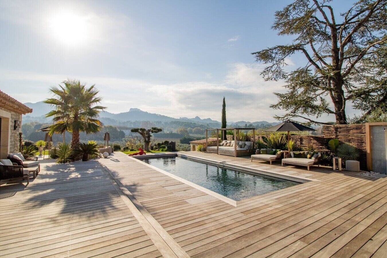 Zona de piscina con cubierta de madera, mobiliario exterior y vista a las montañas.