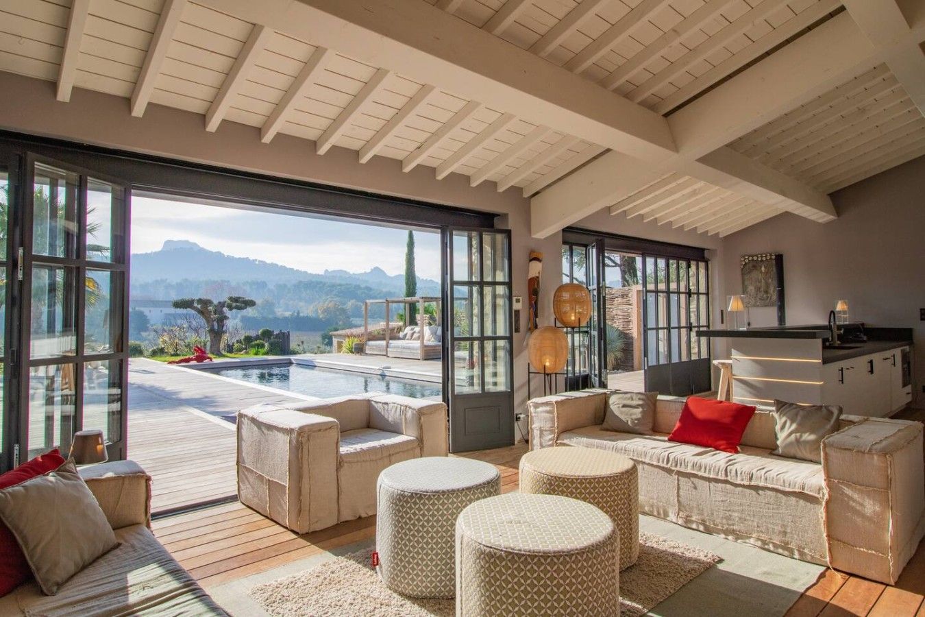 A spacious living area with a large window overlooking a pool and mountains. There is a kitchen area with modern appliances and a dining table.