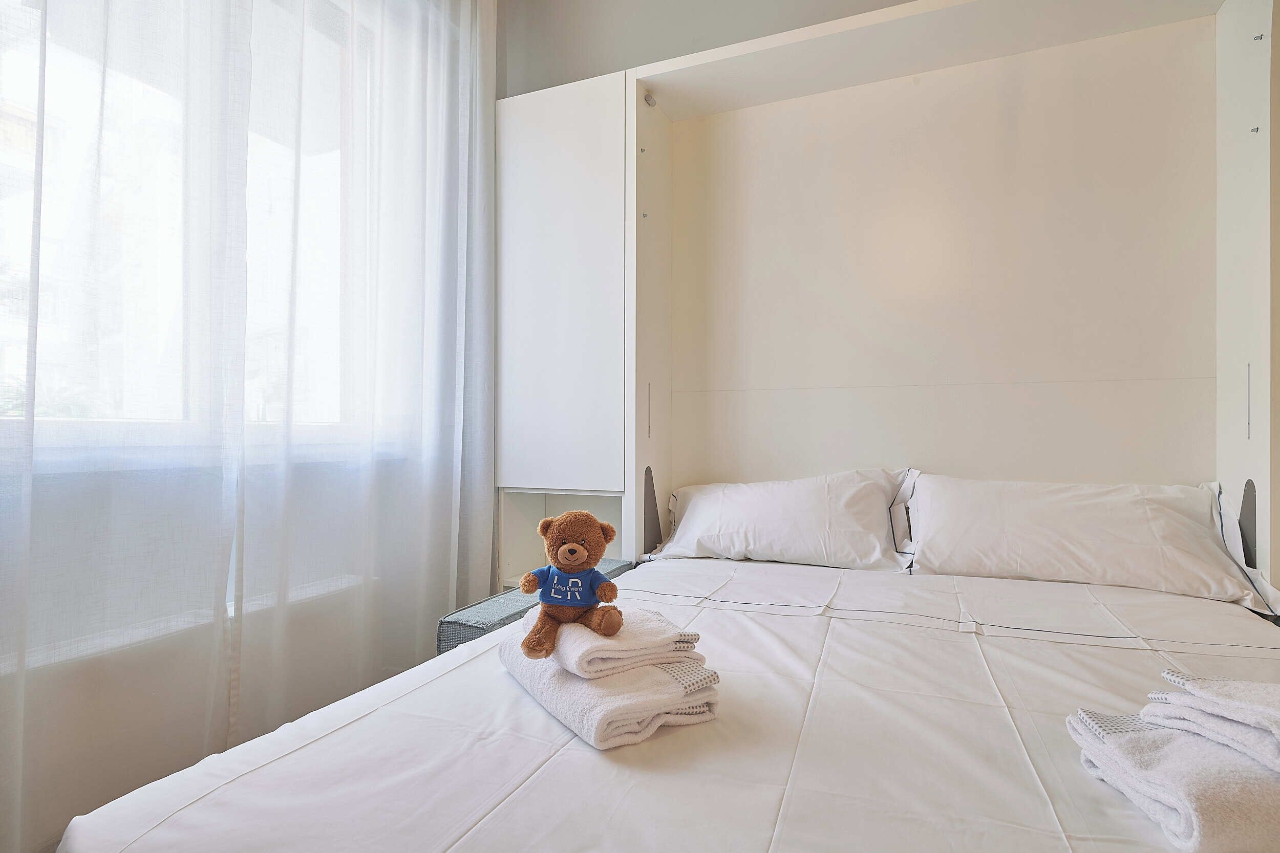 Ein Hotelzimmer mit einem Bett, einem Teddybären und einem Fenster mit durchsichtigen Vorhängen.