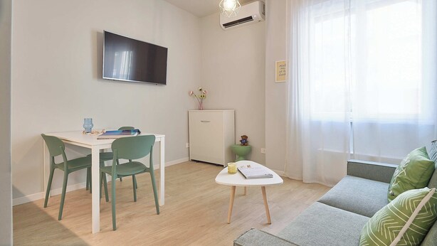 Ett kompakt vardagsrum med en matplats, en soffa, en tv och ett fönster med tunna, genomskinliga gardiner.