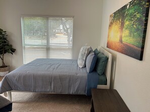 A bedroom with a bed, a window with blinds, a wall-mounted painting, and a potted plant.