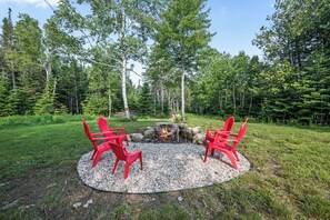A circular seating area with a fire pit surrounded by red chairs, set in a forested area with trees and grass. - Hildy's Hideaway | NEW! Sleeps 10 | Direct Snowmobile Access | Near Saddleback & Downtown (Rangeley)