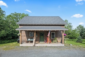 A two-story wooden house with a porch, a green door, and a small American flag. - Hildy's Hideaway | NEW! Sleeps 10 | Direct Snowmobile Access | Near Saddleback & Downtown (Rangeley)