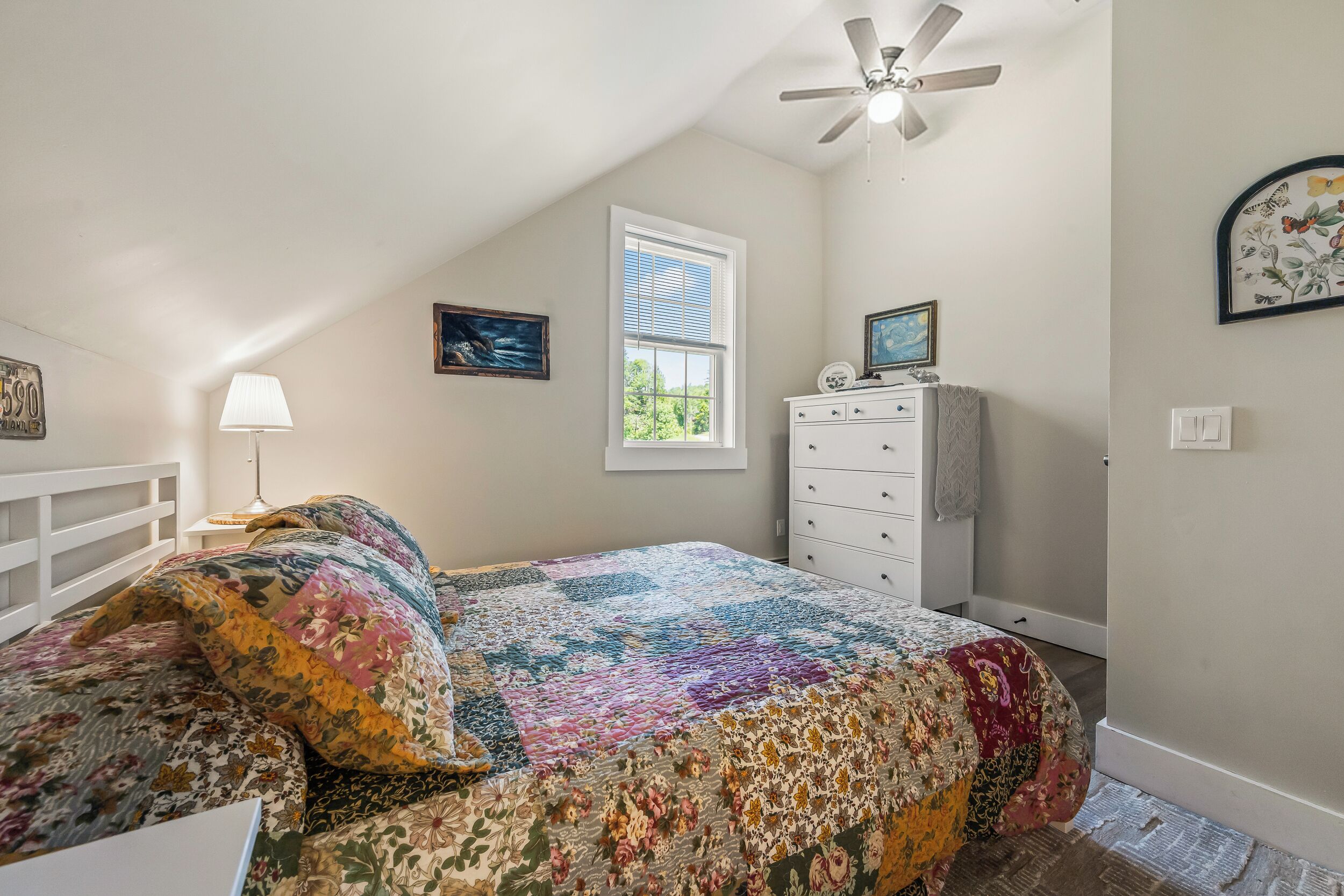 A bedroom with a sloped ceiling, a bed with a colorful quilt, a nightstand with a lamp, a dresser, and a window with blinds.