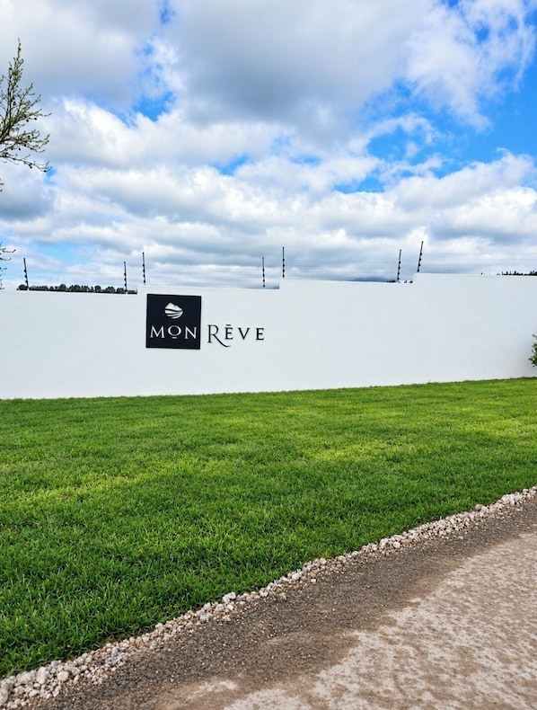 Un letrero con la inscripción 'MON RÉVE' sobre una pared blanca, un campo cubierto de hierba y un cielo nublado.