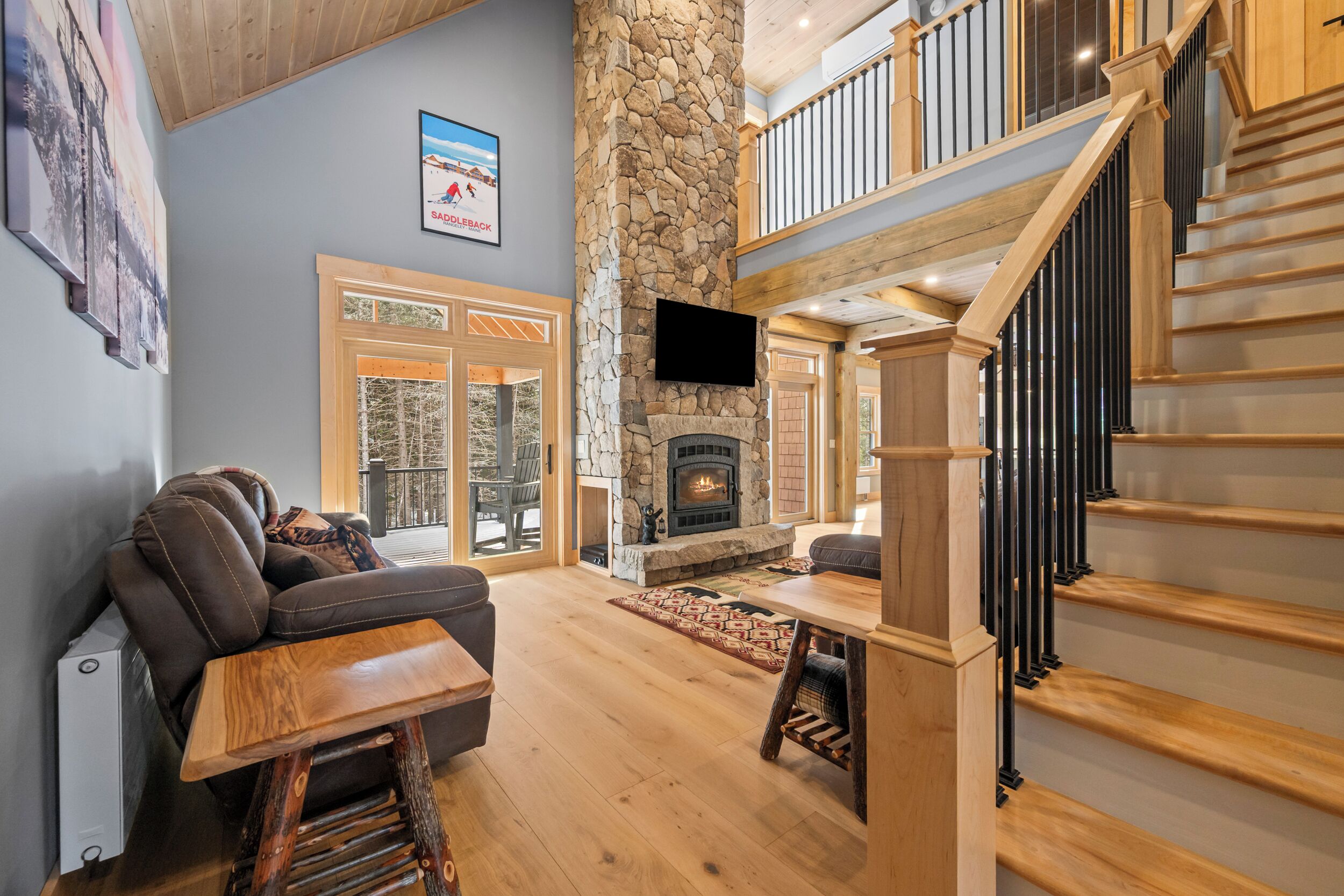 A spacious living area with a stone fireplace, a wooden staircase, and a large window overlooking a deck.