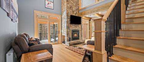 A spacious living area with a stone fireplace, a wooden staircase, and a large window overlooking a deck.