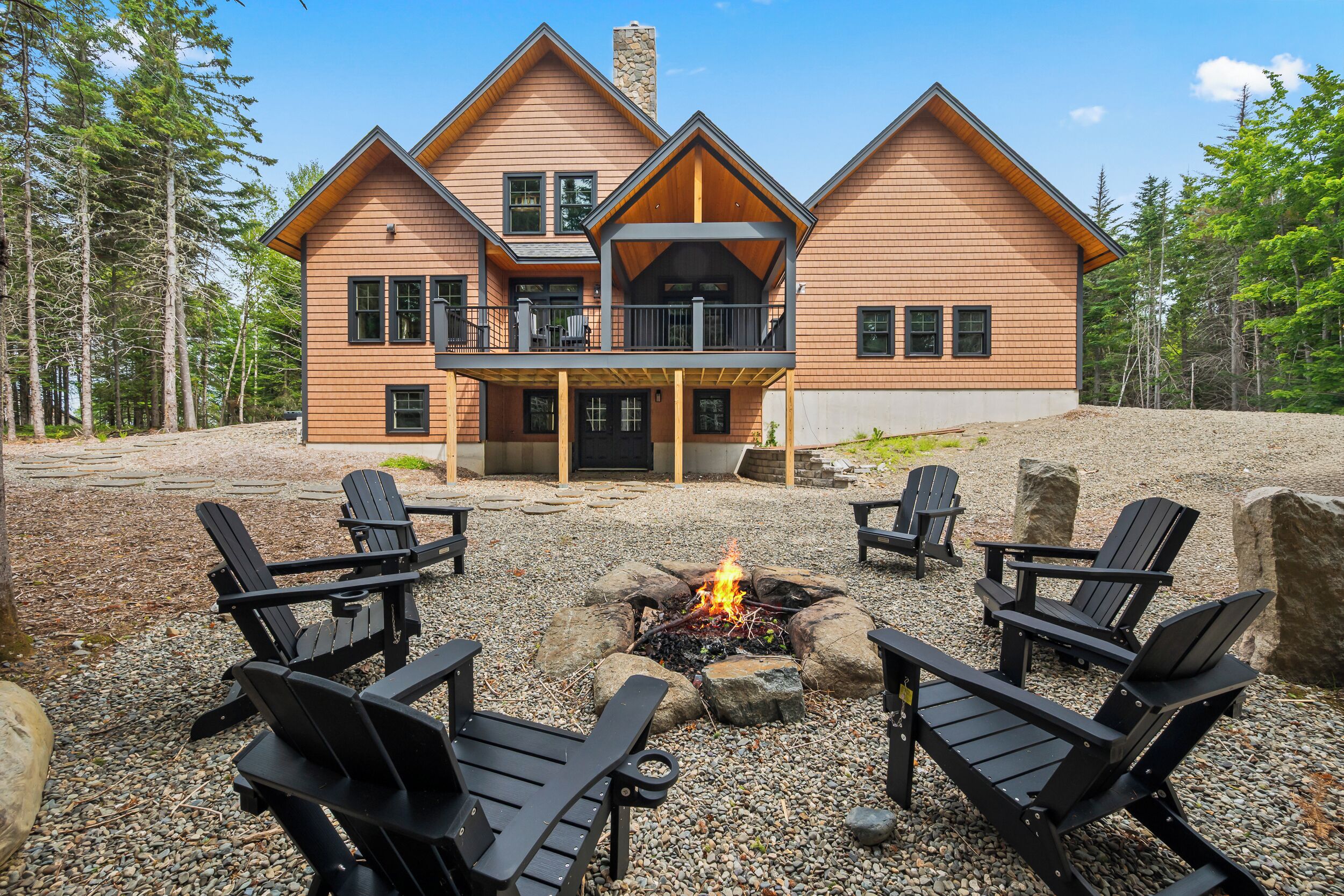 A two-story log cabin with a balcony, surrounded by a gravel area with Adirondack chairs and a fire pit.