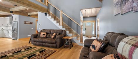 A spacious living room with a high ceiling, wooden staircase, and a kitchen in the background.
