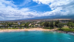 A coastal resort with a sandy beach, clear turquoise waters, and a backdrop of hills and buildings. - Kihei Condo Across from Kamaole Beach Near Dining and Shopping (Kihei)