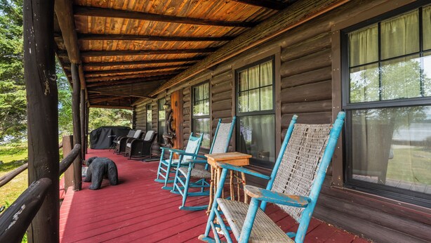 A covered porch with wooden flooring, a row of rocking chairs, and a view of the surrounding forest. - The Boston | Waterfront on Mooselookmegtunic Lake | Pet Friendly (Rangeley)