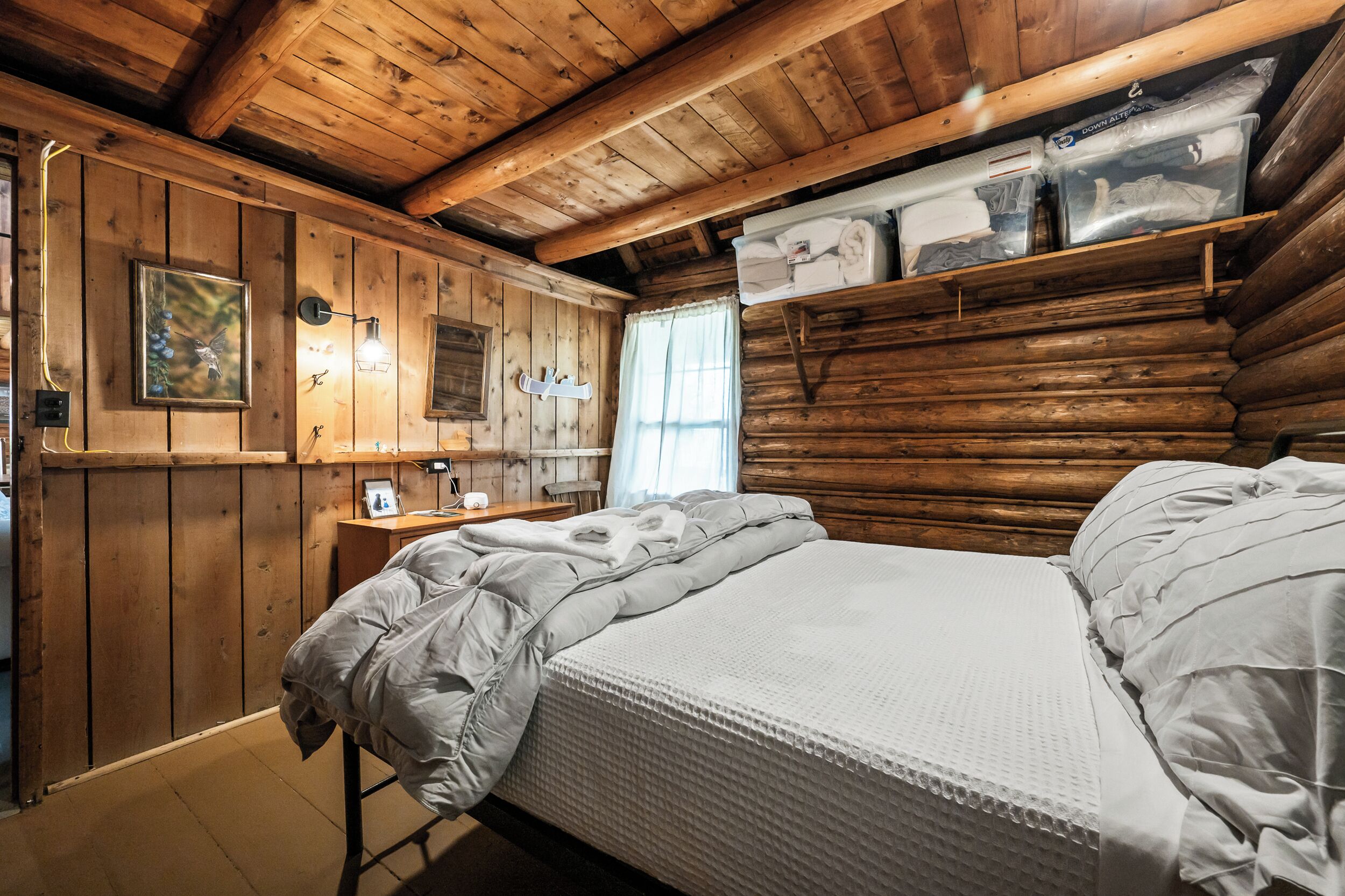 A log cabin room with a bed, wooden walls, a window, and a shelf with folded towels.