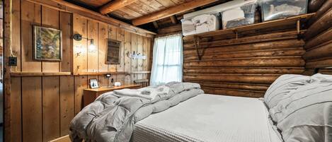 A log cabin room with a bed, wooden walls, a window, and a shelf with folded towels.