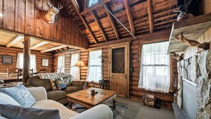A log cabin interior with a wooden ceiling, a fireplace, a sofa, a coffee table, and a stone wall. - The Boston | Waterfront on Mooselookmegtunic Lake | Pet Friendly (Rangeley)