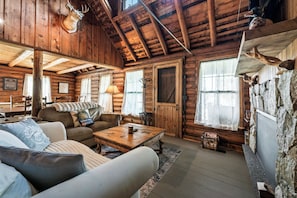 A log cabin interior with a wooden ceiling, a fireplace, a sofa, a coffee table, and a stone wall. - The Boston | Waterfront on Mooselookmegtunic Lake | Pet Friendly (Rangeley)