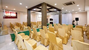 Un salón de conferencias con hileras de sillas tapizadas en tela color beige, un escenario con alfombra verde y un telón de fondo blanco.