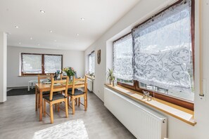 A modern dining area with a wooden table and chairs, a window with decorative curtains, and a radiator.