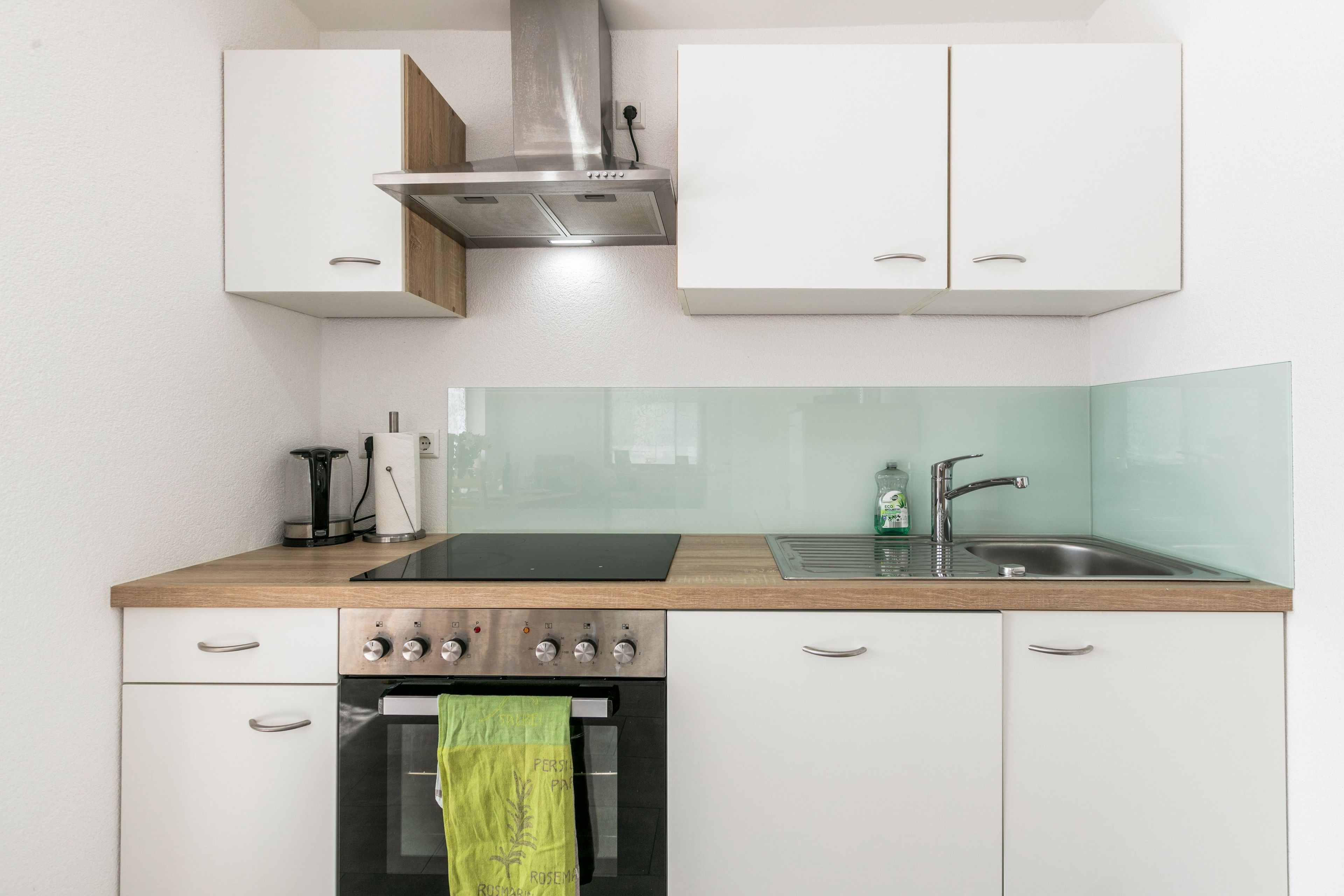 Une cuisine moderne avec des armoires blanches, une hotte de cuisinière en acier inoxydable, un four encastré, un évier avec un robinet et un pare-feu en verre.