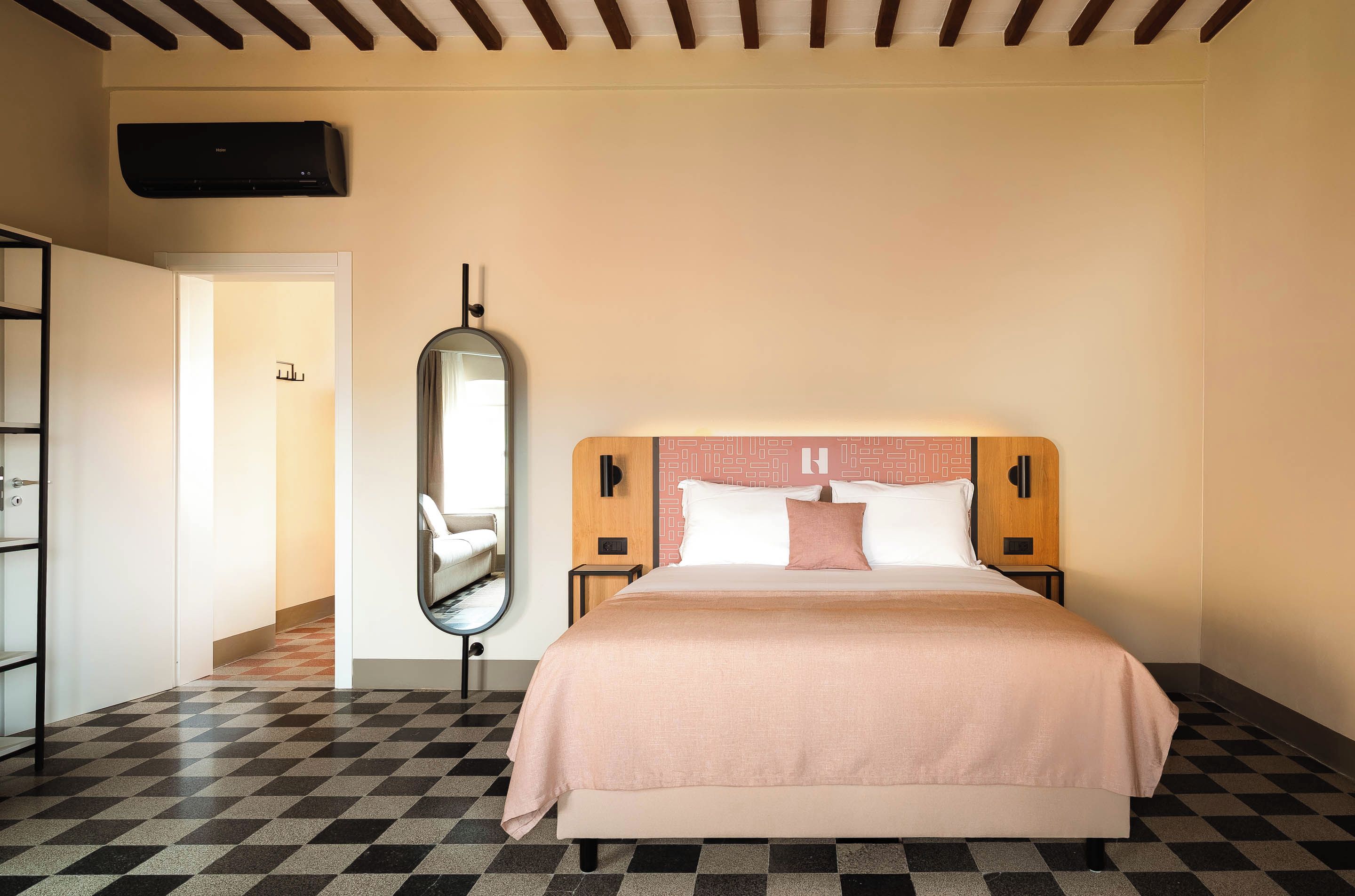 A hotel room with a checkered floor, a bed with a pink bedspread, a large mirror, and a wall-mounted air conditioning unit.