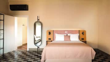 A hotel room with a checkered floor, a bed with a pink bedspread, a large mirror, and a wall-mounted air conditioning unit.