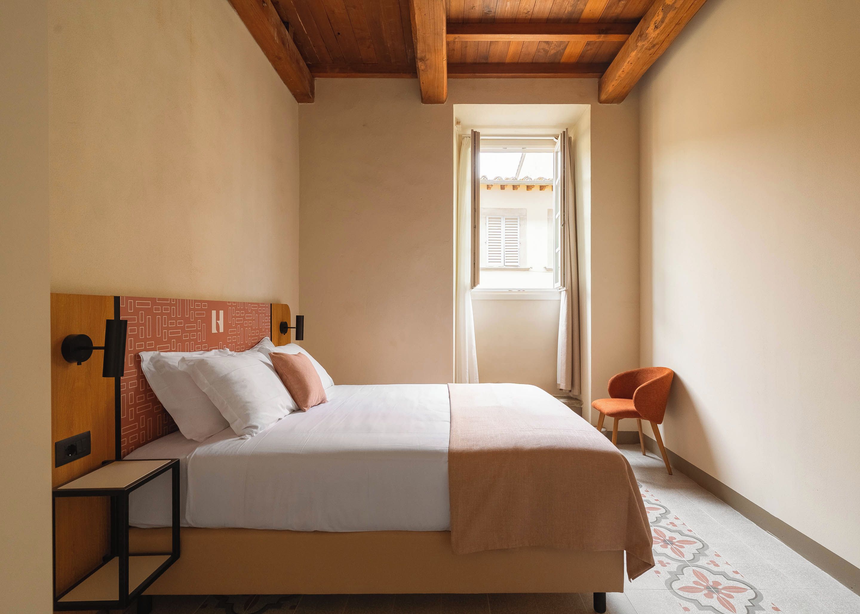Een slaapkamer met een bed, nachtkastje, stoel en raam met gordijnen.