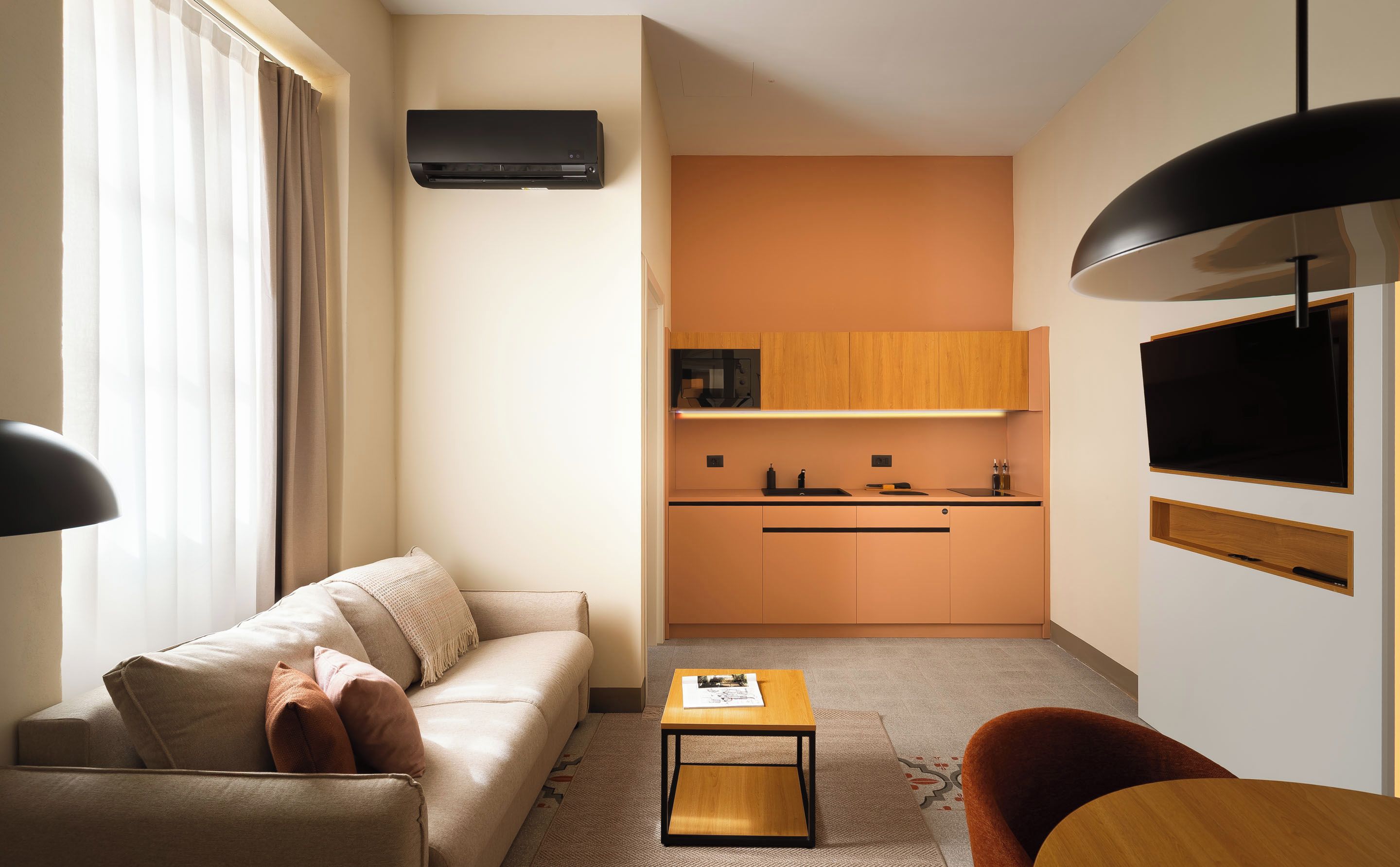 A compact living space with a sofa, a small table, a kitchen area, and a TV mounted on the wall.