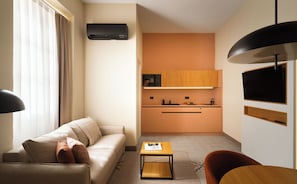 A compact living space with a sofa, a small table, a kitchen area, and a TV mounted on the wall.
