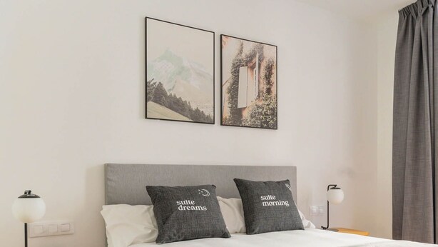 Una cama bien hecha con dos almohadas, una mesita de noche con lámpara y dos cuadros en la pared.