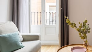 Un salón con sofá, una mesa redonda de madera con un cuenco, y vista a un balcón a través de un ventanal.