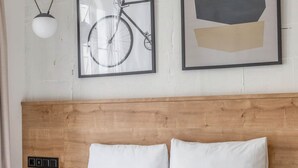 Una habitación de hotel con cabecera de madera, ropa de cama blanca, una toalla enrollada y dos cuadros en la pared.